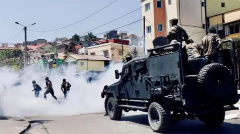 Reuters Riot Police in a heavily armoured bear-cat kind of vehicle drive behind three protesters who are running away through tear gas smoke.