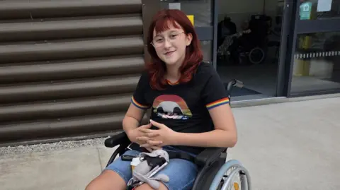 Family handout Wren is smiling into the camera. We see her in her manual wheelchair. She is wearing a blacktop with rainbow edging on the short sleeves and blue denim shorts. 
Wren has dark red hair and is wearing glasses. 
