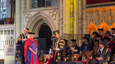 Gillian stands on stage in her cap and gown on graduation day. In front of her, on stage, are other graduates in gowns and wearing caps. They appear to be in a cathedral.