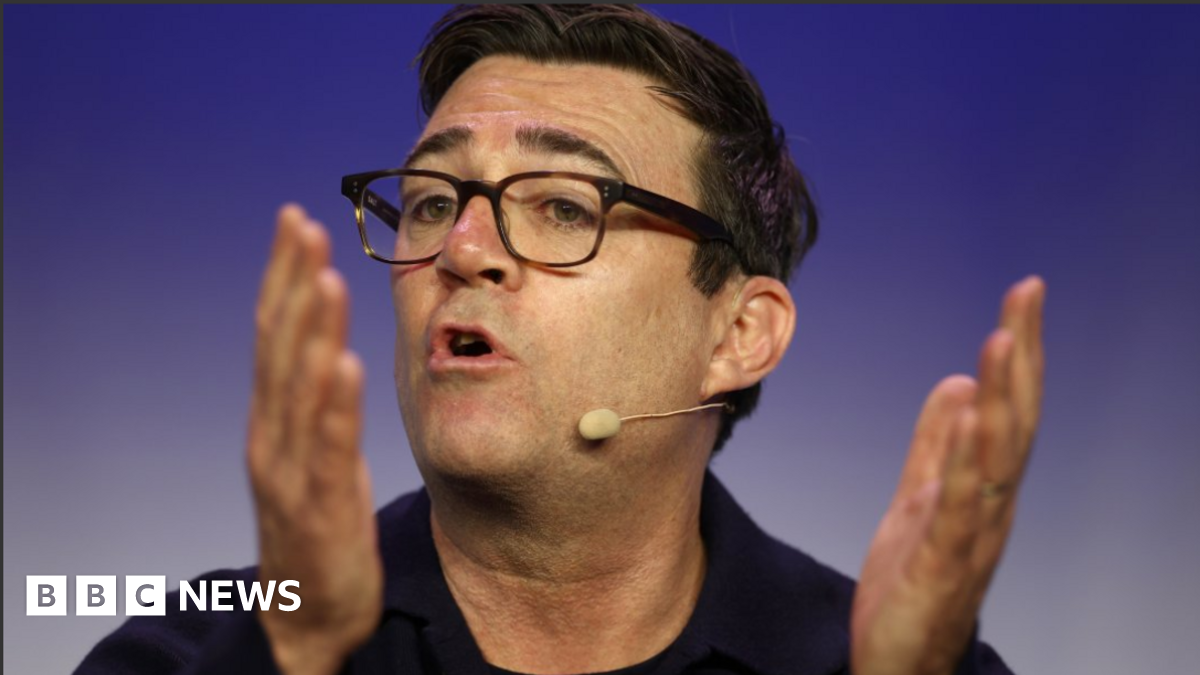 Mayor of Greater Manchester Andy Burnham speaking wearing a headset mic. He has dark brown hair, cropped medium short, and wears dark tortoiseshell glasses.