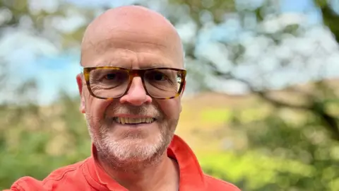 Colm Martin Colm Martin, in a dark orange T-shirt, smiling into the camera. He is outdoors, with the background blurred