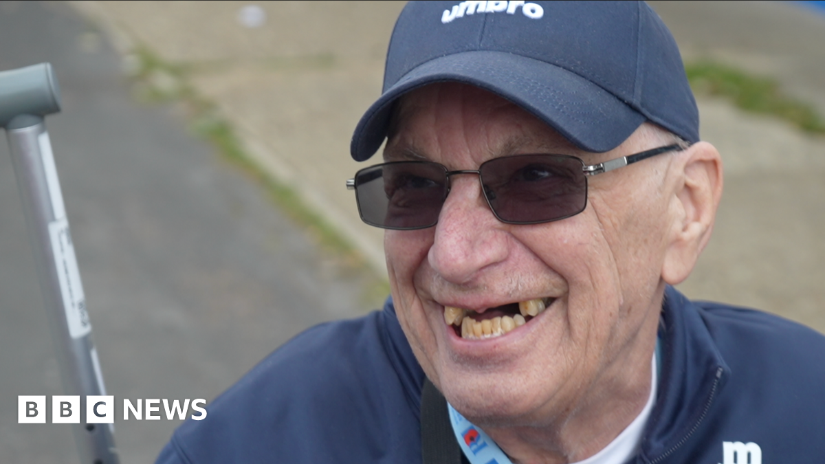 A head shot of 76-year-old John Haddock. He is wearing dark glasses, a navy hat, and a navy top. He is looking past the camera and smiling.