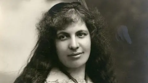 Pitt Rivers Museum Black and white image of a Māori woman wearing a traditional feather cloak with a feather in her hair. She is wearing a traditional piece of pounamu round her neck.