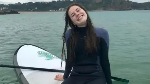 Megan McDonagh Lily sat on a paddle board in a wetsuit smiling at the camera. Her long dark hair is wet from the sea. 