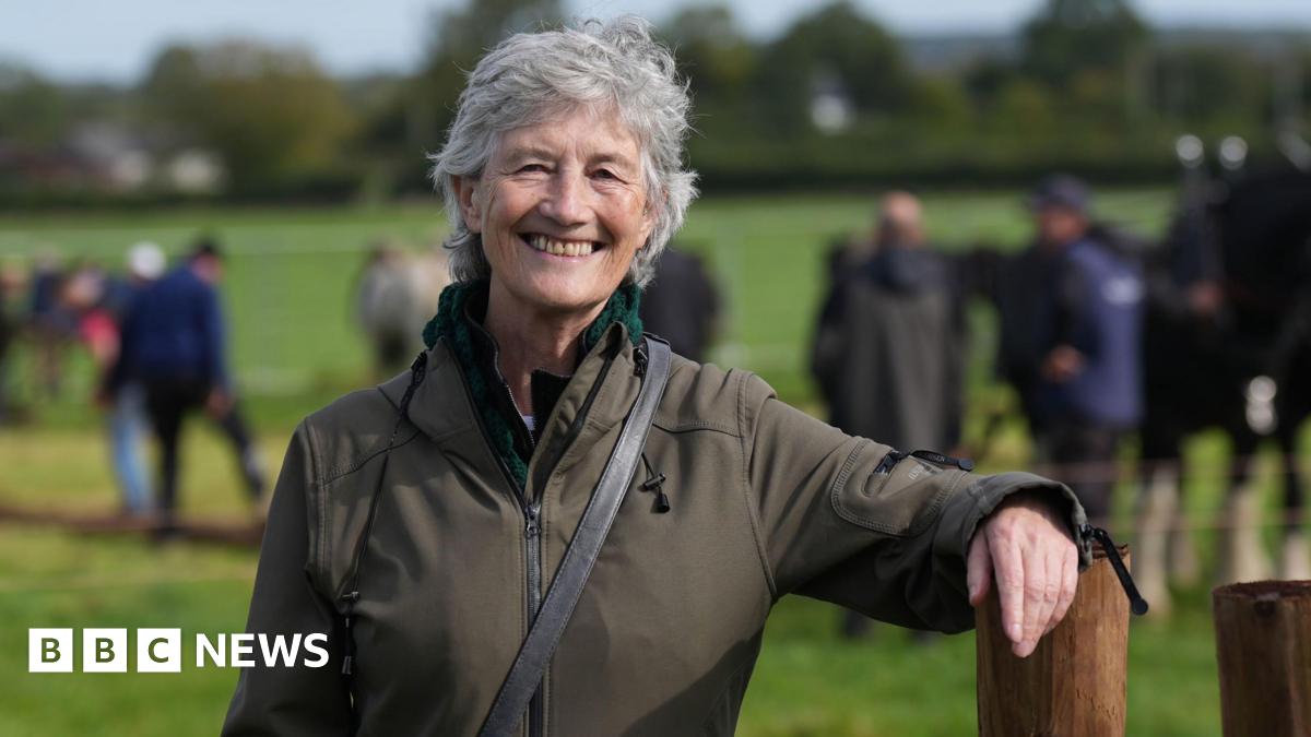 Catherine Connolly wears a green jacket. She has short grey hair and is smiling. She is standing in a field and leaning her arm on a post.