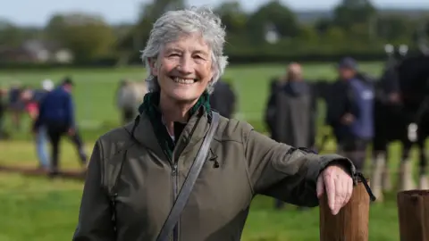 PA Media Catherine Connolly wears a green jacket. She has short grey hair and is smiling. She is standing in a field and leaning her arm on a post. 