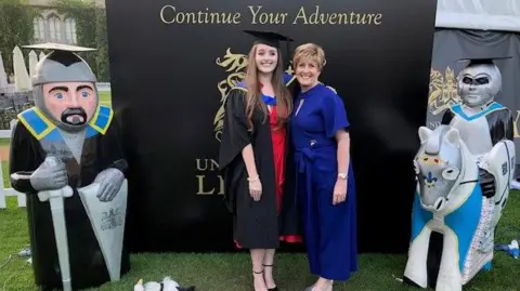 University of Lincoln Grace in a cap and gown stands smiling with her mother, who wears a blue dress. They are standing outside in front of a black backdrop, with the wording printed "continue your adventure".