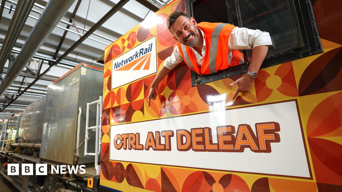 A man leans out the window of a train and points at the name painted on the side of it. He is wearing an orange high-vis jacket and the train is painted with orange and green leaves.