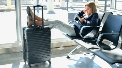 Getty Images A blonde woman sits in an airport chair with her feet up on her wheeled suitcase, looking at her phone.