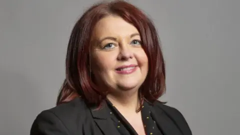 UK Parliament Headshot of Liverpool Wavertree MP Paula Barker. She has shoulder length  dark, auburn hair and is wearing a black jacket and a black top with small yellow spots. 