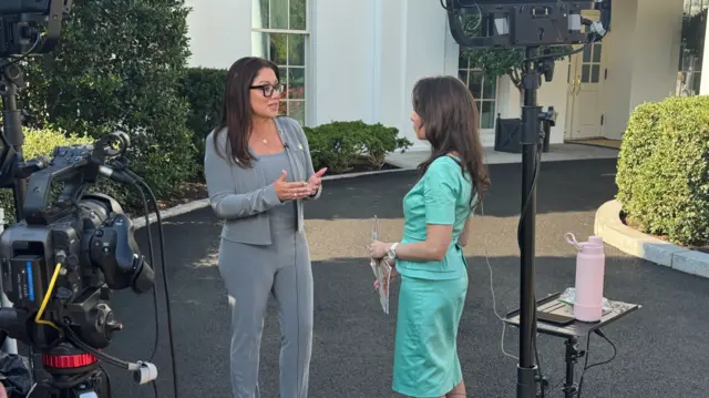 Two women speak in front of a camera and microphones outside the White House