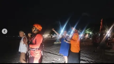 Denpasar Search and Rescue A member of search and rescue wears an orange helmet on a beach. It is dark and he is with three other people