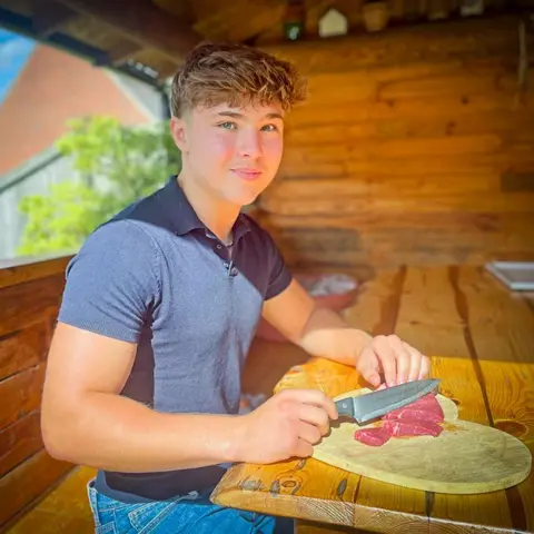 Ruth Clegg, BBC Nat Walney, who has brown hair and blue eyes, sat in a wooden cabin, sat on a wooden bench and eating raw steak from a plate on a wooden table. He is wearing a navy T-shirt and looking at the camera slightly smiling.