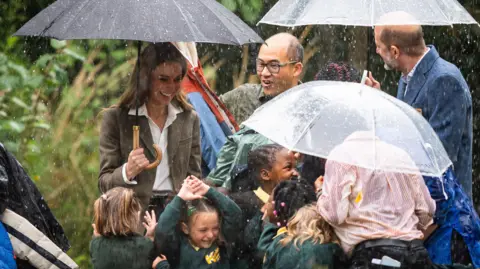 PA Media Four adults including Catherine, who is holding one of the three umbrellas in the rain and five young schoolchildren. 