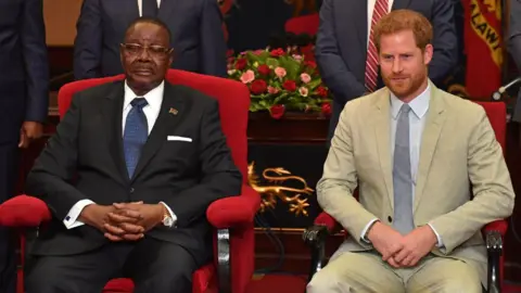 WireImage via Getty Images Peter Mutharika, wearing a black suit, sits next Prince Harry, who wears a beige suit. They are both seated in red chairs  and figures stand behind them.