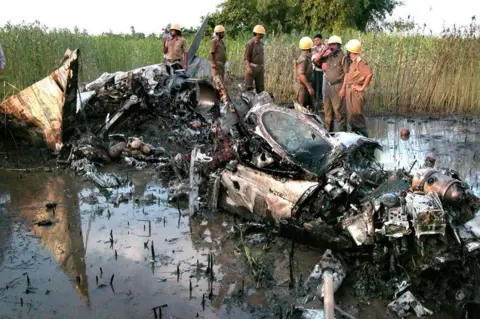 AFP via Getty Images Indian firefighters stand beside the wreckage of a MiG-21 military plane that caught fire before crashing, 15 July 2002, in Phansidawa near Siliguri in the state of West Bengal.