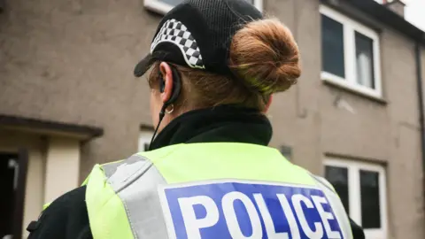 the back of a female police officers with orange hair tied into a bun, peaking through the back of a police cap. He is wearing a hivs vest which says police on it