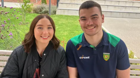 BBC Eimhear O'Neill and Luke MacDaibheid sitting on a bench beside each other. They are both looking at the camera and smiling. Ms O'Neill is wearing a black coat and she has brown hair. Mr MacDaibheid is wearing a green and navy Donegal GAA polo. He has brown hair. 