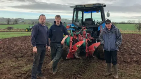 Jack Wright Three men stand in front of a blue tractor with plouging equipment attached. They are in a field, with grey skies and hills behind. There is another tractor in the distance.