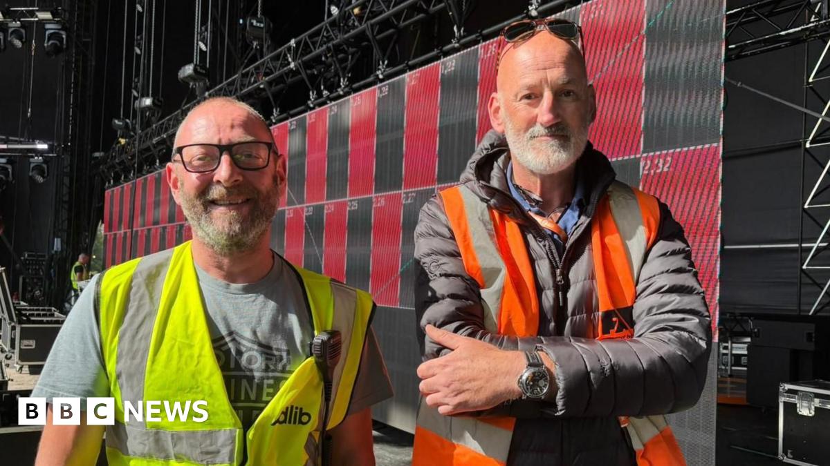 An outdoor music stage with a Radio 2 banner on it and TV screens on the sides. It is being set up, with various workers in hi-vis jackets on stage.