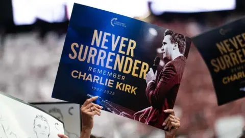 Getty Images Woman's hands holding placard that says 'Never Surrender, Remember Charlie Kirk' with a pic of Kirk holding a microphone.