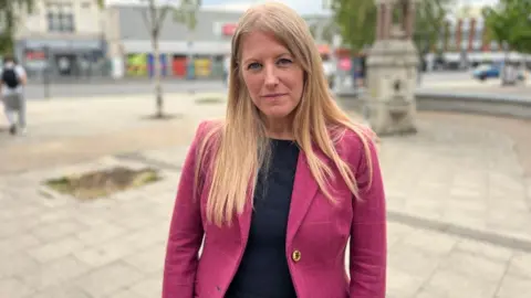 A woman with long blonde hair, in a pink blazer, stands in an empty town square, looking at the camera.