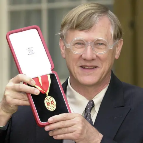 PA Media Sir Nicholas Grimshaw is holding an open box with a medal on a ribbon to the camera as he smiles. The medal is a gold oval shape with red patterning that forms a crown. The ribbon is red and gold striped. Sir Nicholas is wearing a smart black jacket, a pale grey waistcoat, a cream shirt and a black and cream spotted tie. He has straight blond and grey hair and is wearing a pair of glasses. Behind him are windows.