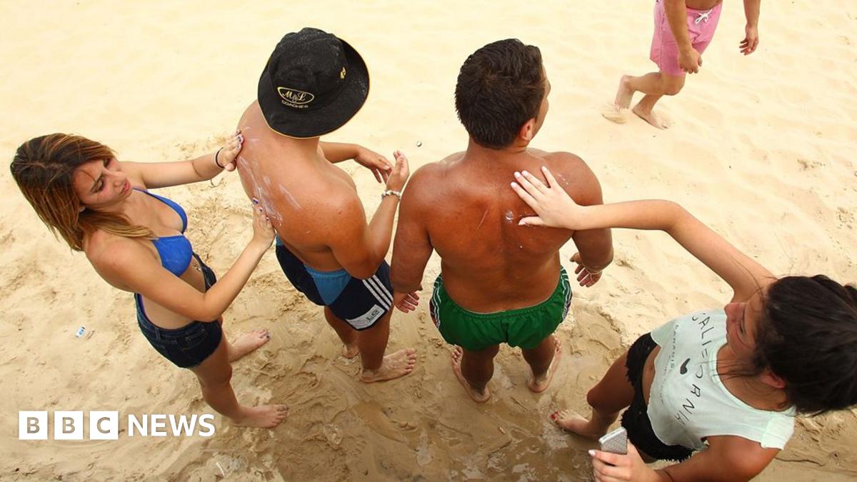 Two women wipe sunscreen on the backs of two men on a beach. The men are wearing swimming shorts while the women are in shorts ad in one case a bikini top and the other a sleeveless white top