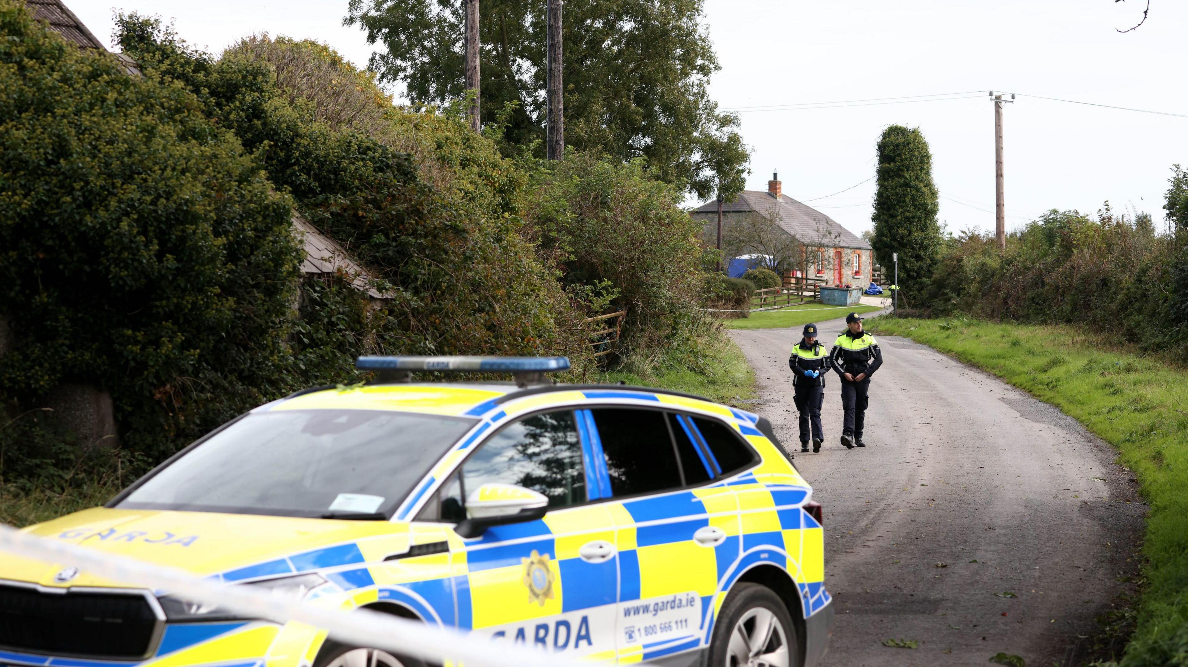 A garda (Irish police) patrol car and two officers manning a cordon on a rural road.  The family's stone cottage is in the background in the distance. 