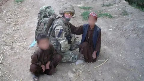 Carole Timone Carol Timone crouching on a dirt track in Afghanistan wearing combat fatigues and a helmet and carrying a large backpack. There are two young boys either side of her wearing traditional Afghan dress. The faces of the two boys have been blurred.