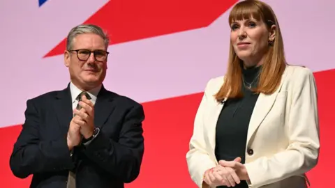 Getty Images Angela Rayner stands on stage at Labour Party conference in September 2025, while Sir Keir Starmer applauds in the background.