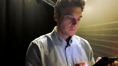 BBC Joe Tidy looking down at a phone. He has short brown hair and is wearing a light blue shirt.