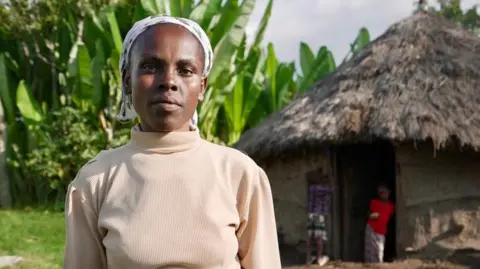 Amensisa Negera / BBC Getenesh Gabiso wearing a beige jumper and headscarf. She is looking at the camera, with a thatched hut and tall plants visible behind her.