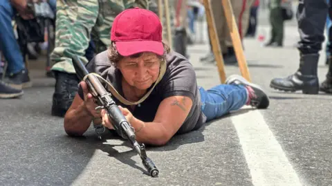 Nicole Kolster/BBC An older woman wearing a red baseball cap struggles to hold up a rifle as she lies on the ground. She is looking at the ground while the rifle also points at the tarmac. In the background there are several people wearing camouflage uniforms and military boots. 