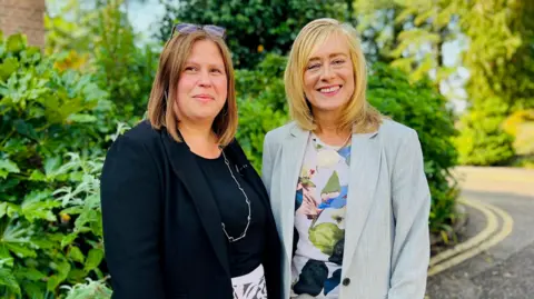 BBC/Branwen Jeffreys Standing outside in front of greenery in the sunshine are the two female Vice Chancellors who have negotiated the merger.  On the left is Professor Georgina Randsley de Moura, the interim Vice Chancellor of the University of Kent wearing a black jacket and top.  On the right is Professor Jane Harrington the Vice Chancellor of the University of Greenwich wearing a pale grey jacket with a patterned floral top. Both women are smiling
