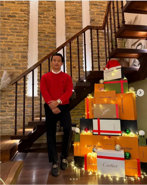 Instagram / sgtthb Saugat Thapa, in a red sweater and black trousers, stands next to a Christmas tree made of boxes of luxury brands 