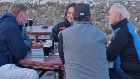 The Beach Bar Sligo A photo is taken of four people sat down at a wooden table outside. 
The centre of a photo is a man with his back turned wearing a grey body warmer with a black baseball cap.
To the right is a man with a blue and black jacket he has grey hair. 
Another man is pictured to the left with a navy raincoat. 
A woman is also pictured with her phone in her hand. 
Some pint glasses filled with dark beer are also sitting on the table. 