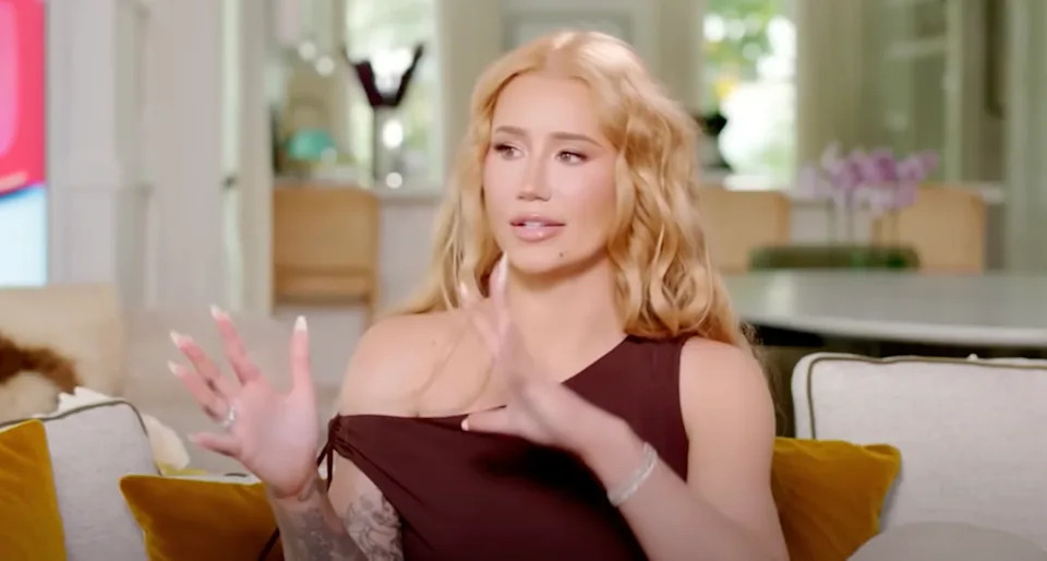 Iggy Azalea sitting on a sofa, gesturing with her hands, in a well-lit room with a modern interior.