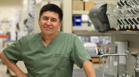 OHSU/Christine Torres Hicks Man in olive-green scrubs in a laboratory with taps and scientific equipment in the background, smiling at the camera.