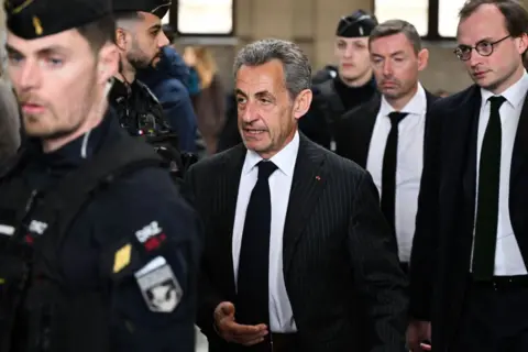 AFP via Getty Image Former President of France Nicolas Sarkozy stands walks as he is flanked by two men, in front and behind him. The two men in front of him is wearing police attire. The two men behind him are wearing matching black suits, white shirts and black ties. The one closest to him is wearing glasses as well. Sarkozy is wearing a black suit with grey pinstripes, paired with a white shirt and black tie. 