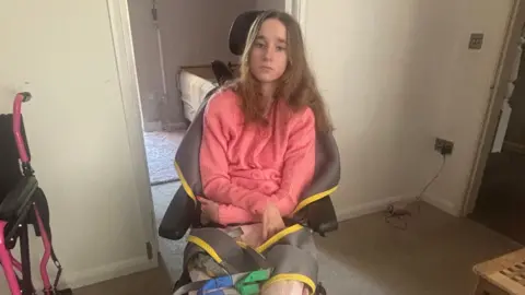 Rachel Cowley A young girl with brown hair in a wheelchair. She is wearing a pink top and has her arms crossed in front of her. There is a pink wheelchair slightly visible on the left side of the image. 