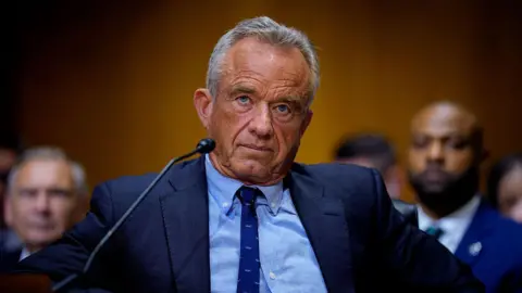 Getty Images RFK Jr photographed at his Senate committee hearing. He is wearing a dark navy blue suit jacket with a slim royal blue tie and a light blue button-up shirt. He is tan, with graying short hair. Behind him are blurred out spectators. 