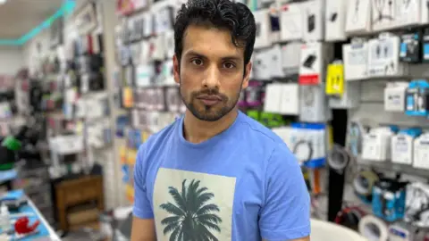 BBC A man with short hair in a blue T-shirt with a palm tree on it looks at the camera without smiling. He is stood inside a phone shop.