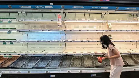 Getty Images A woman stands with a bag of onions in front of a bare supermarket shelf