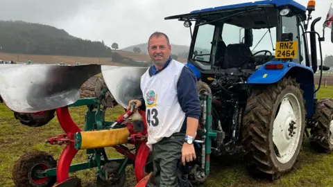 Jack Wright A man stands in front of a blue tractor with plouging equipment attached. He is in a field, with grey skies and hills behind. 