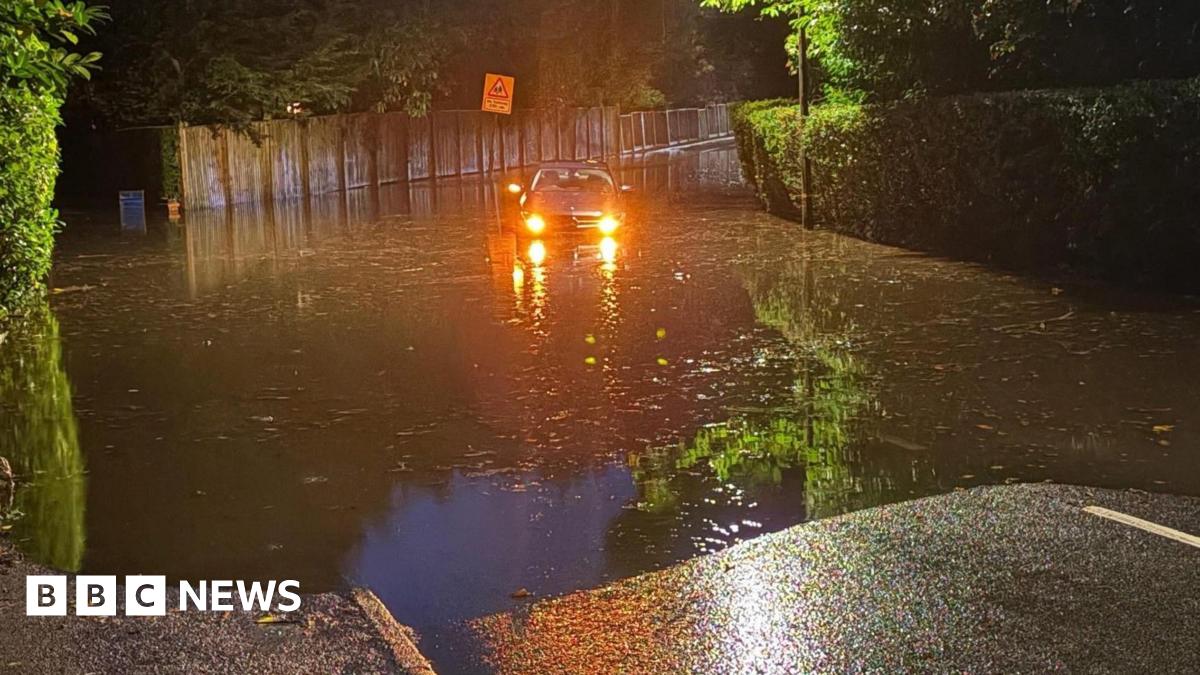 A car, with its headlights on, struggles through water in a flooded street