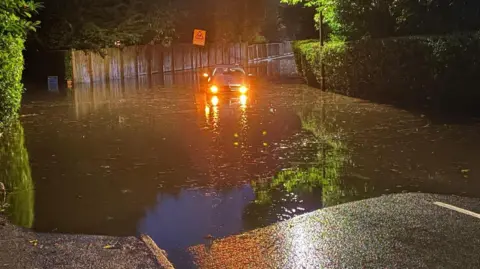 BBC A car, with its headlights on, struggles through water in a flooded street