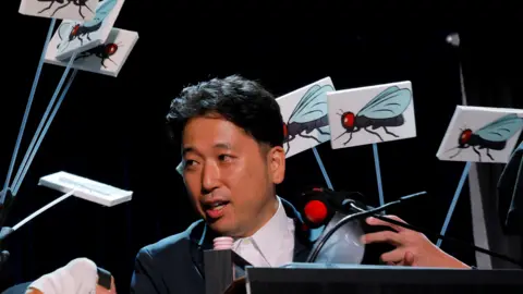 Reuters Researcher Tomoki Kojima of Japan accepts the Ig Nobel Prize for Biology, wearing a dark blazer and white shirt while surrounded by fake flies attached to sticks.