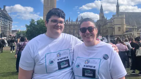 BBC/Kate McGough Tracey wearing sunglasses on the right of frame, hugs her 14 year old son Rowan on the left of frame. Both are wearing white T-shirts with slogans supporting the campaign 'Fight for Ordinary' - a campaign by parents of children with SEND to retain EHCPs during SEND reform. They are both smiling and standing in parliament square with Big Ben in the background. 