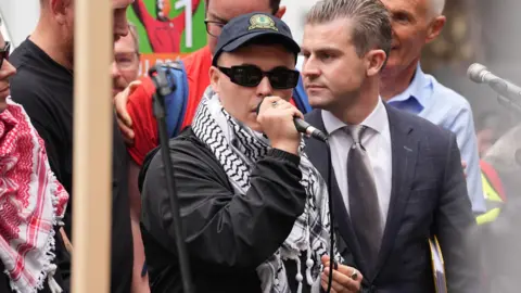 PA Media Liam Óg Ó hAnnaigh, centre, speaking into a microphone. He is wearing a black tracksuit top, a white and black scarf, black sunglasses and a navy hat. He is surrounded by a crowd of people. A man in a dark grey suit, white shirt and grey tie is standing to the right of him. 